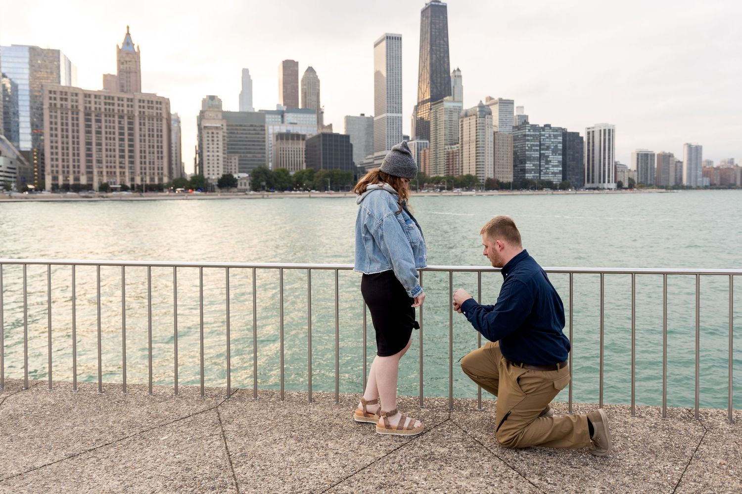 Man proposing on one knee to girlfriend by waterfront with Chicago city skyline in background.