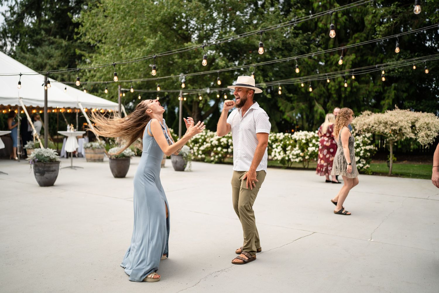 Guests dancing at outdoor wedding reception with string lights and tent in background.