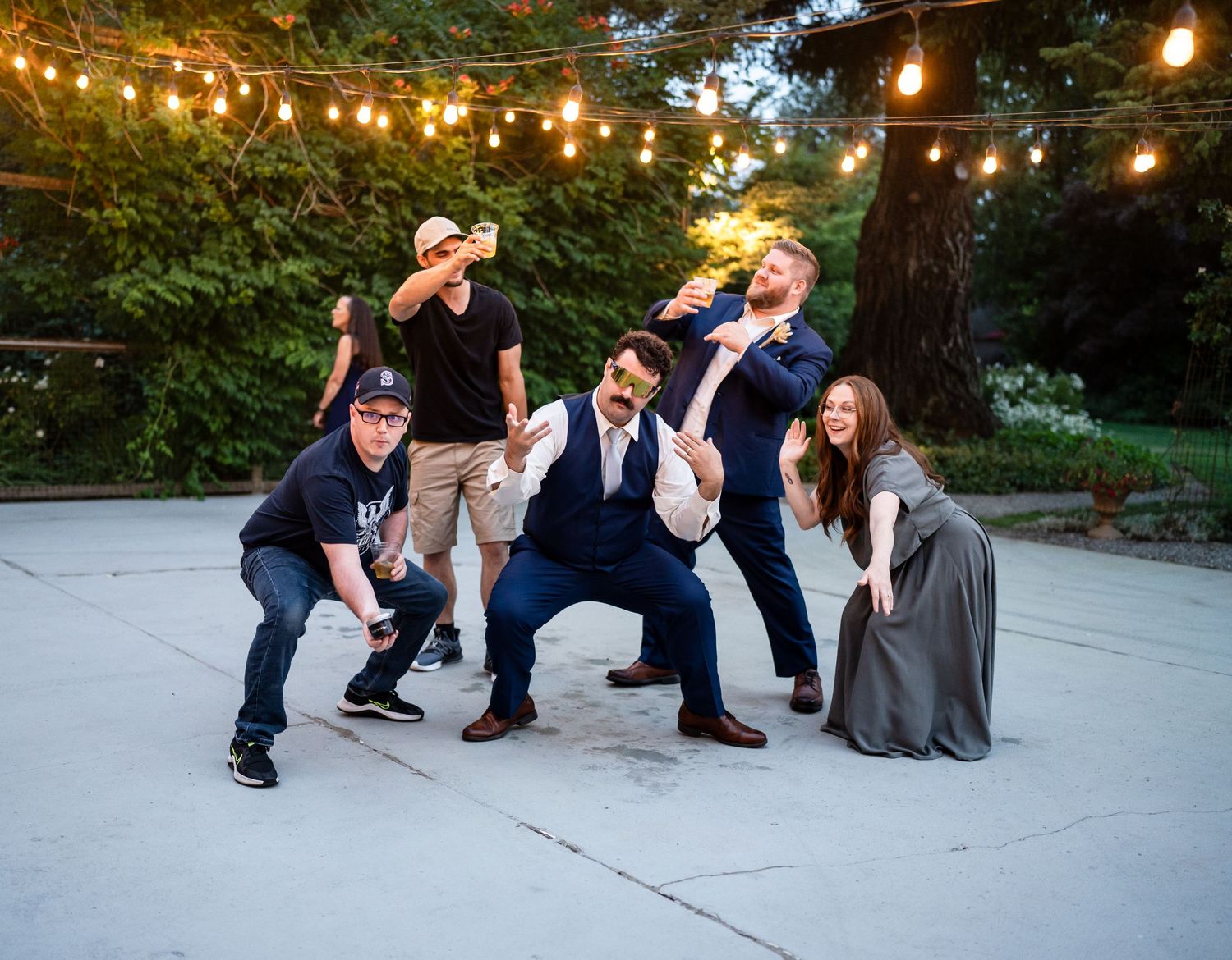 Wedding party poses for fun group photo under string lights at outdoor reception.
