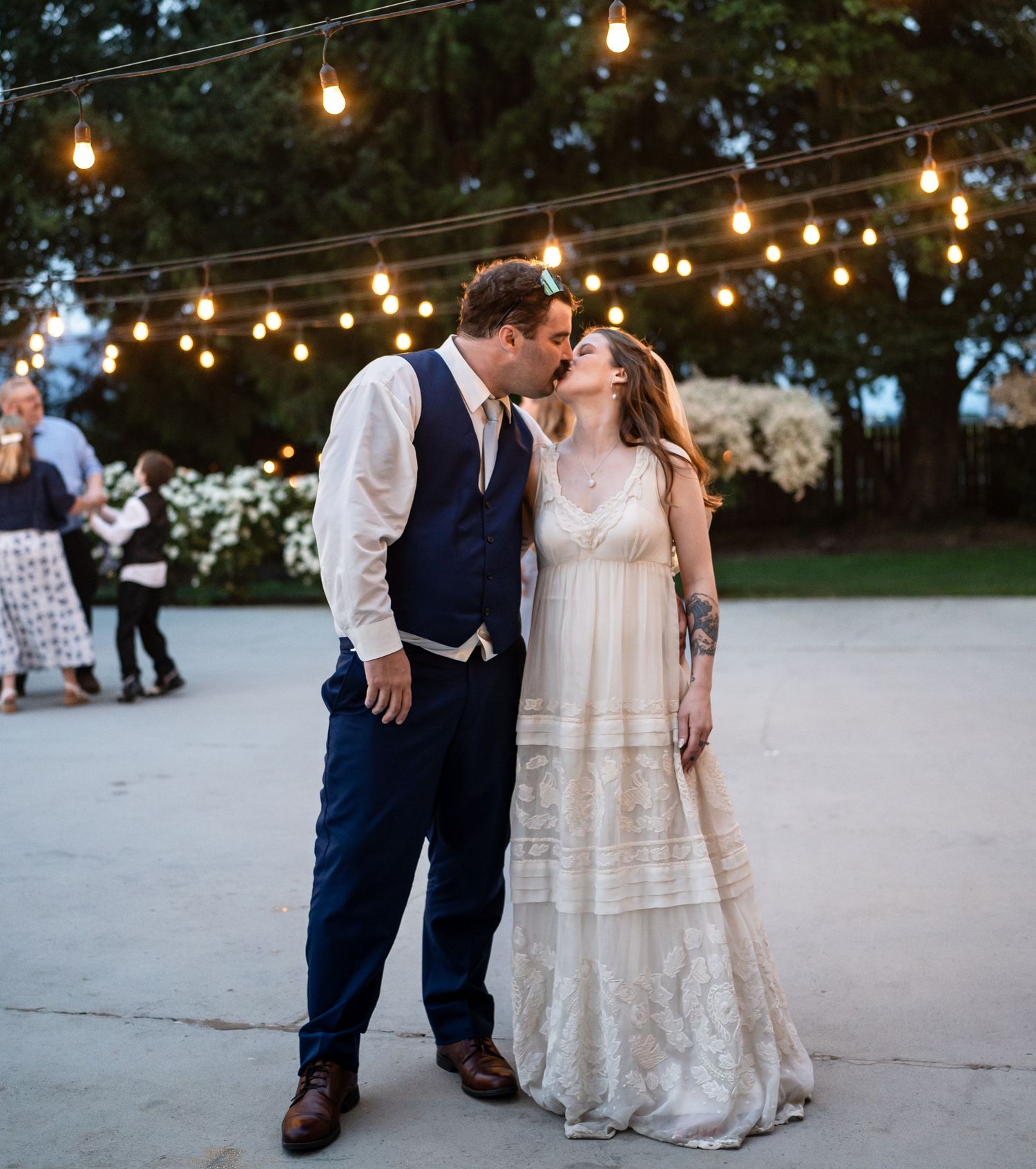 Couple shares romantic moment under outdoor string lights at twilight wedding celebration.