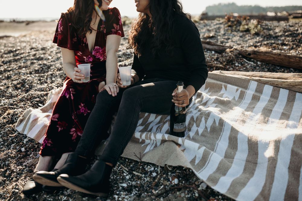 Two people sit together on a striped blanket on a rocky beach at sunset.