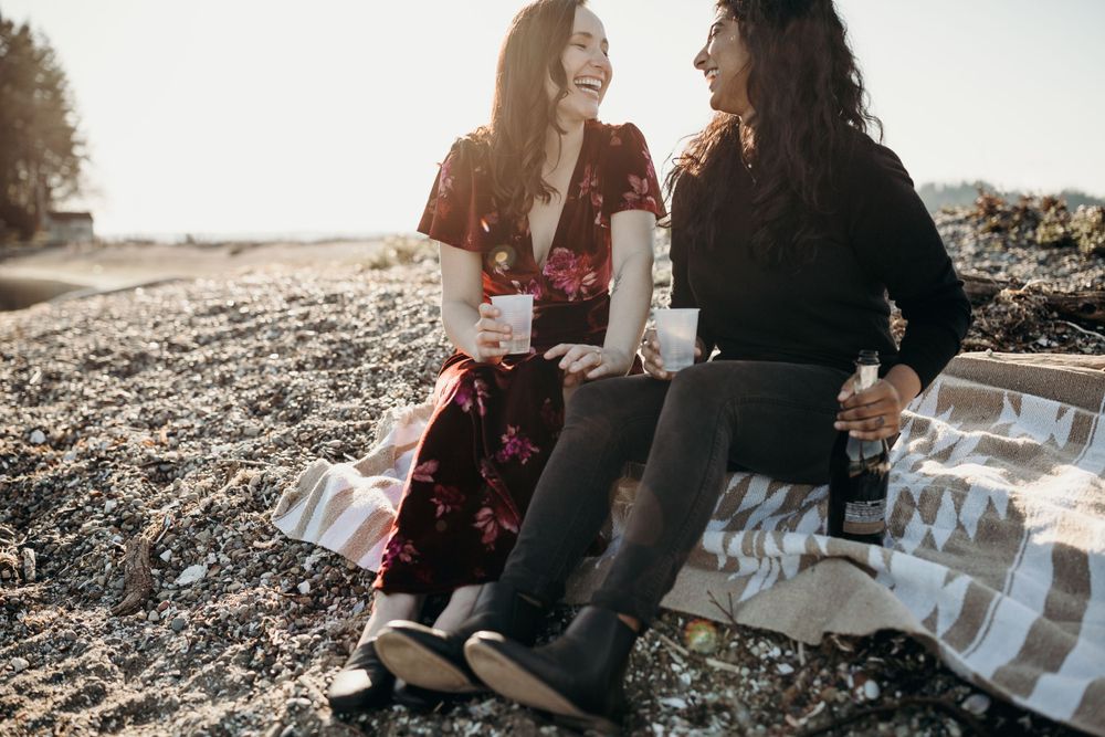 Two people share a laugh while sitting on a blanket at the beach.