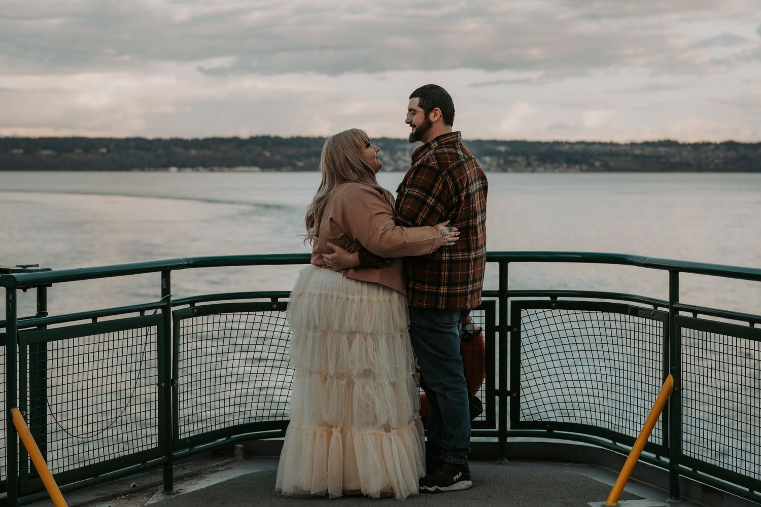Couple in a tulle skirt and plaid shirt hugging on a ferry deck with a cloudy waterfront behind them.