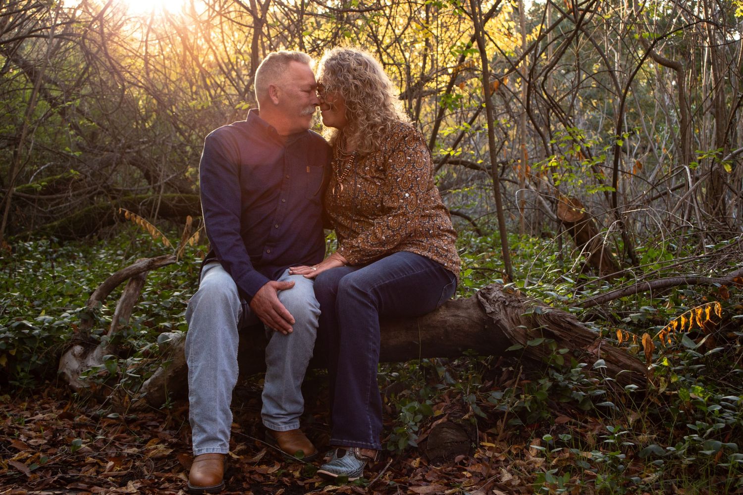 Couple sitting on a log in the woods, smiling, nose-to-nose, with the sun shining behind them.