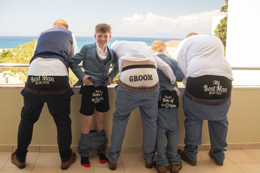 Groomsmen showing embroidered boxer shorts with 'Groom' text while overlooking a scenic coastal view.