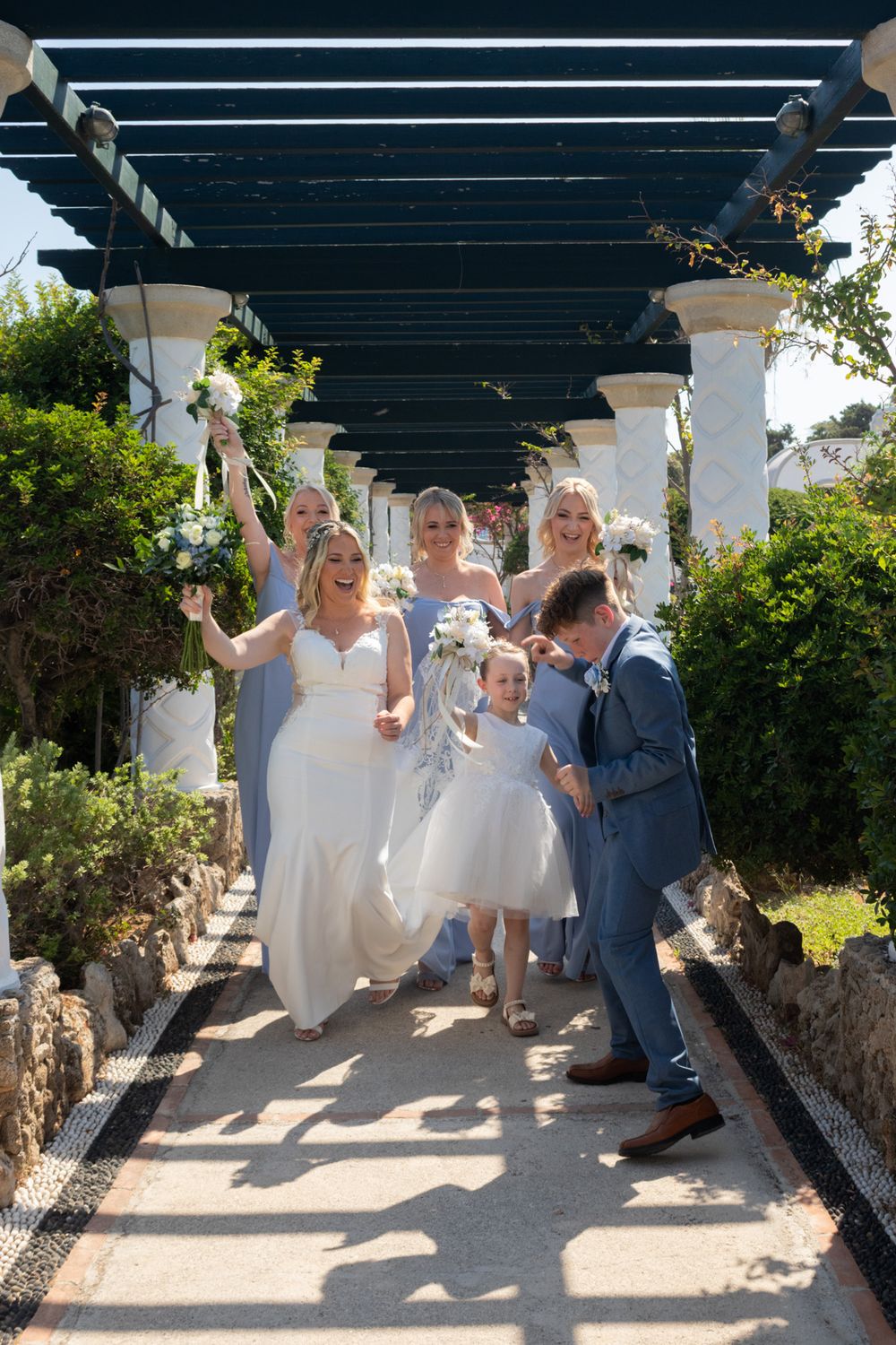 Wedding party walking under a pergola decorated with greenery on a sunny day.