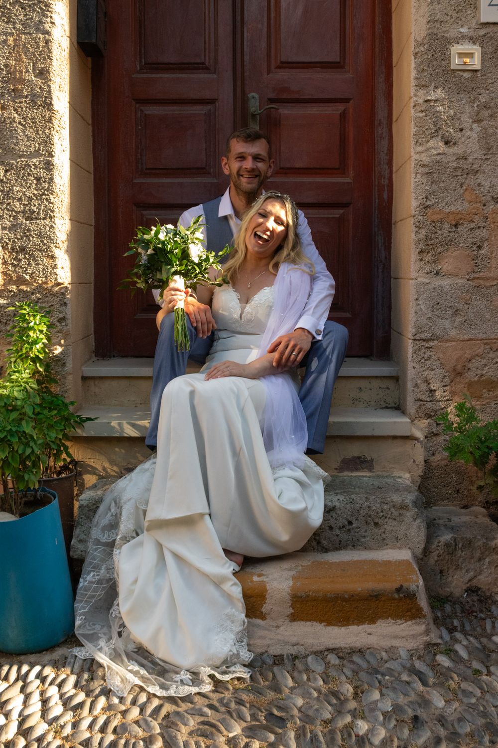 Newlyweds posing on rustic stone steps in front of wooden door with floral bouquet.