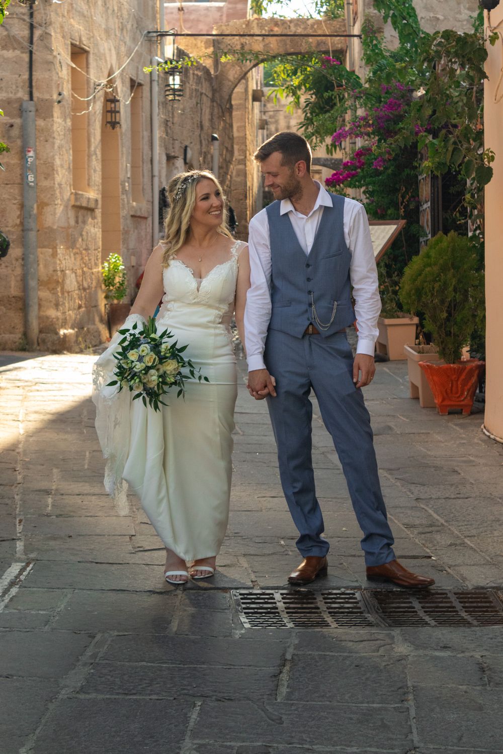 Couple walking hand in hand through Mediterranean street with stone buildings and flowers.