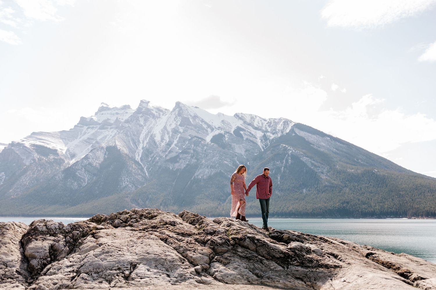 Two figures stand on rocky outcrop with dramatic mountain vista behind.