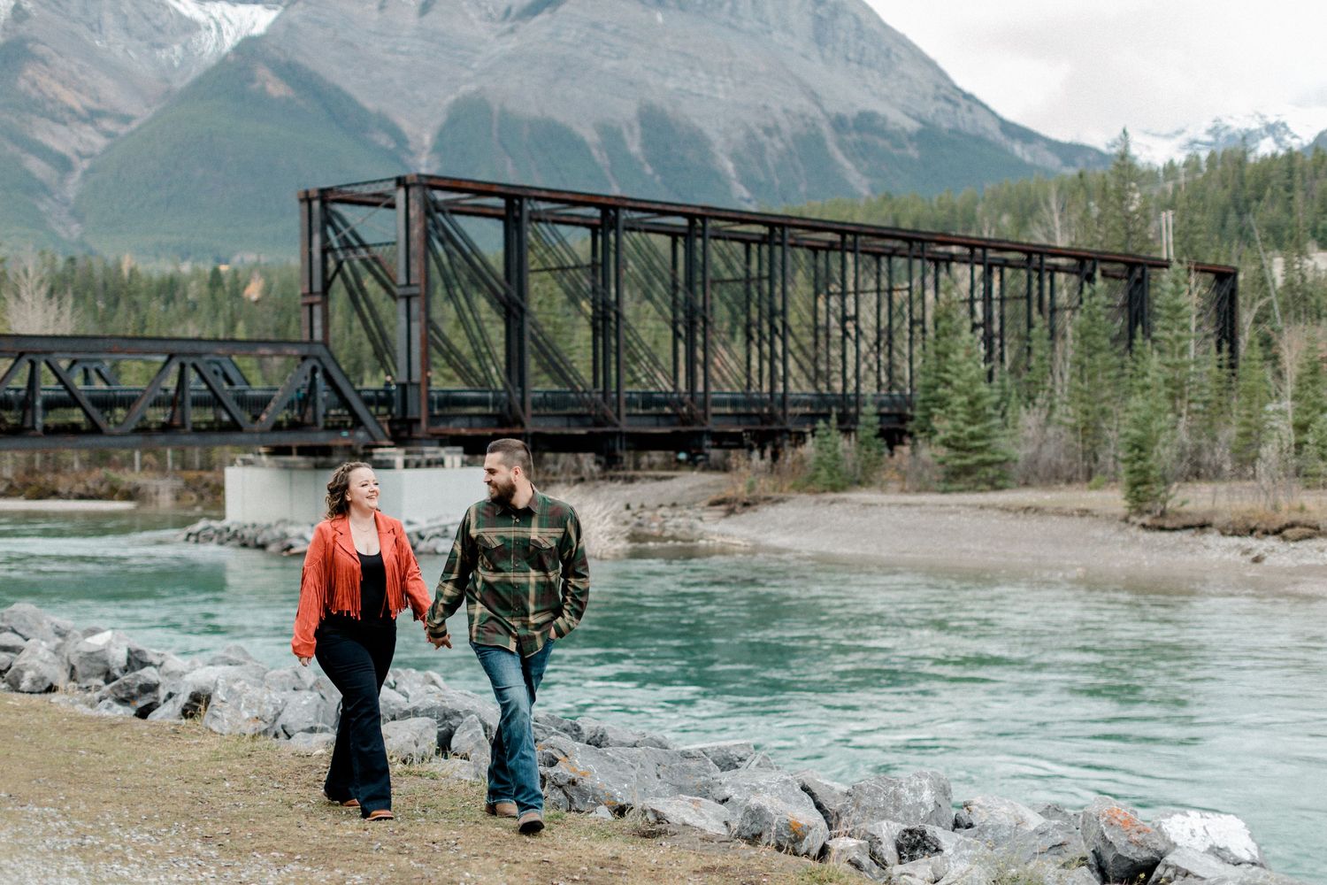 A couple walks along a riverbank with a steel bridge and mountain backdrop in the Canadian Rockies.
