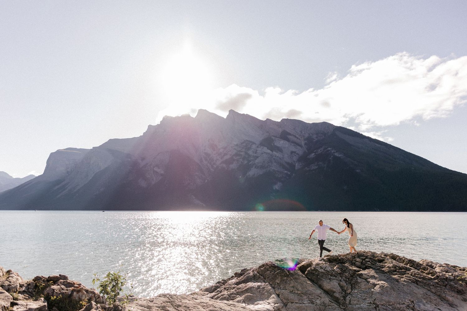 Two people stand on rocks by a mountain lake during sunset with dramatic mountain backdrop.