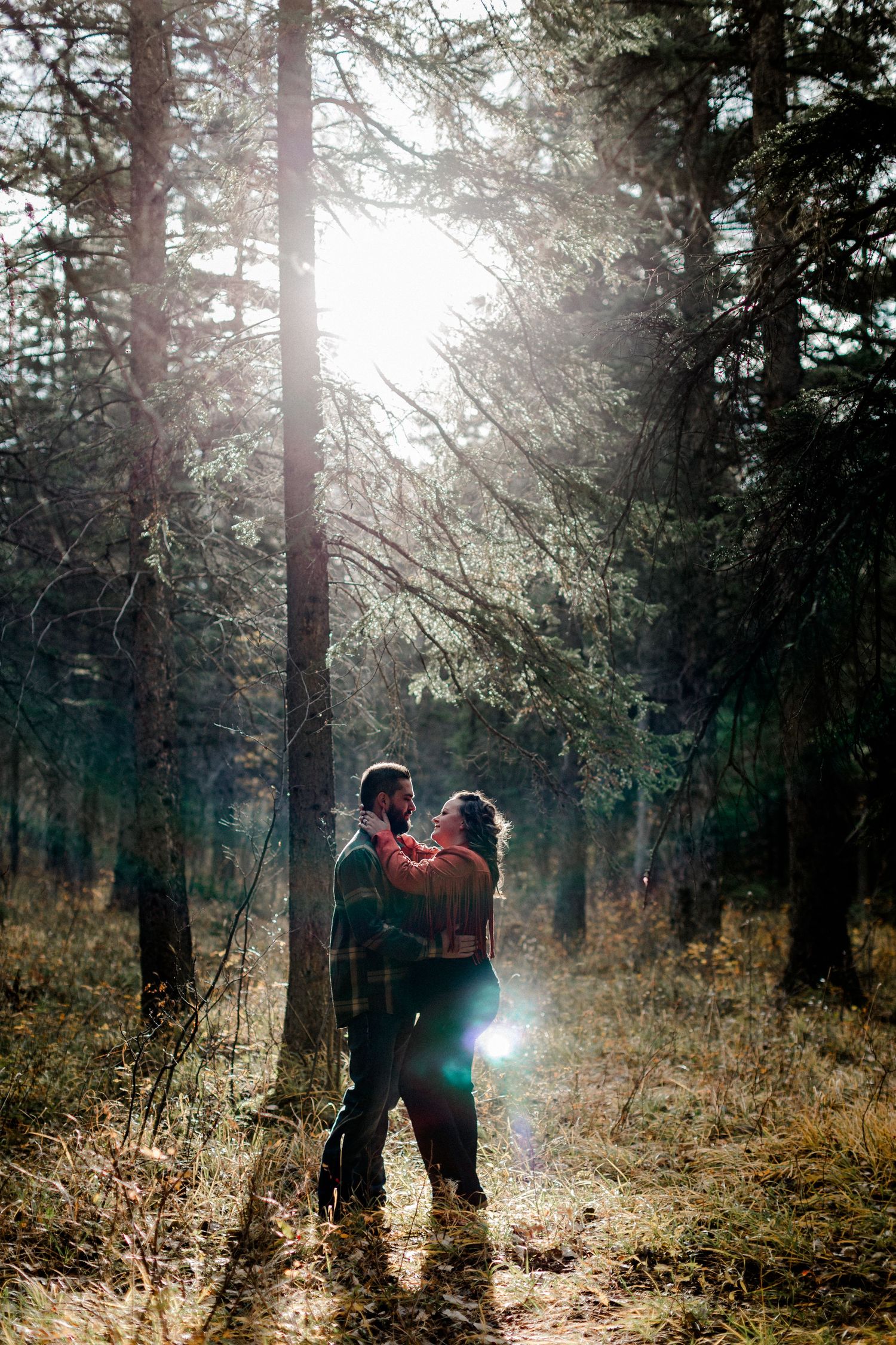 Silhouettes embrace in a sunlit forest with rays streaming through evergreen trees.