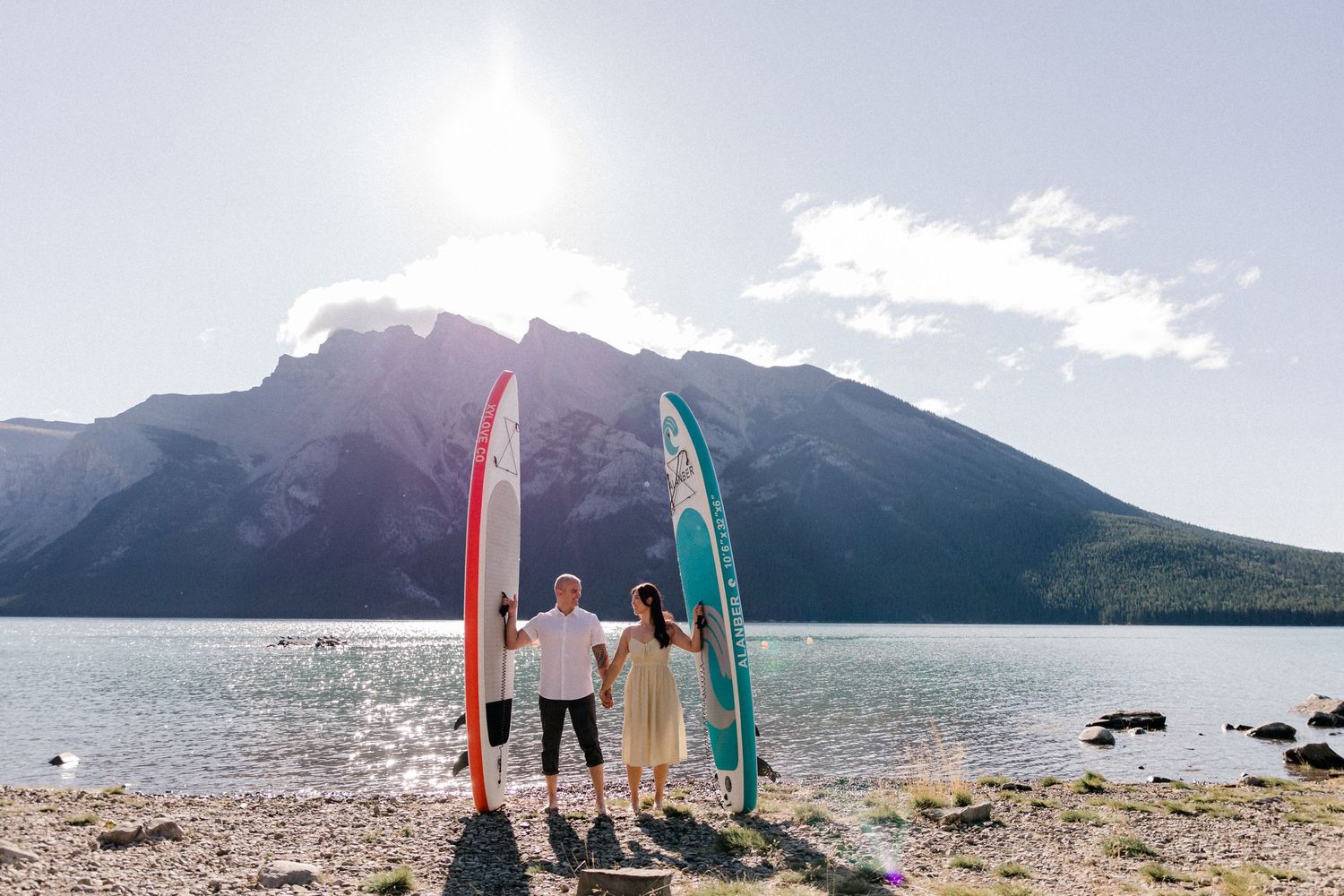 Three people stand on a rocky beach holding colorful surfboards against a mountain backdrop.