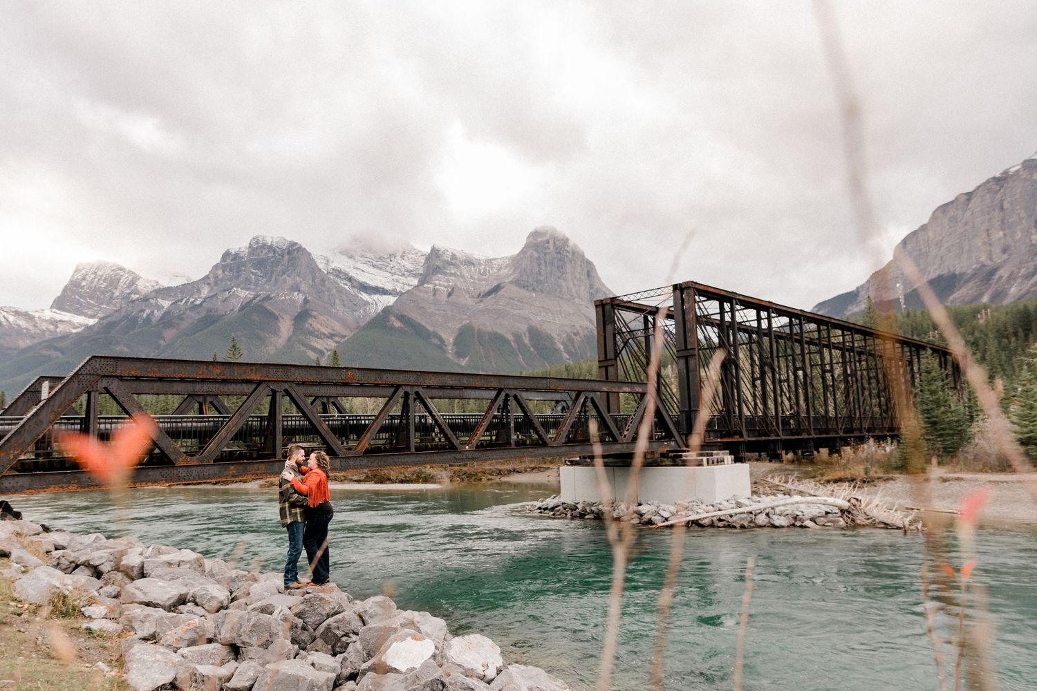 Historic steel bridge spans turquoise river with majestic mountain backdrop.