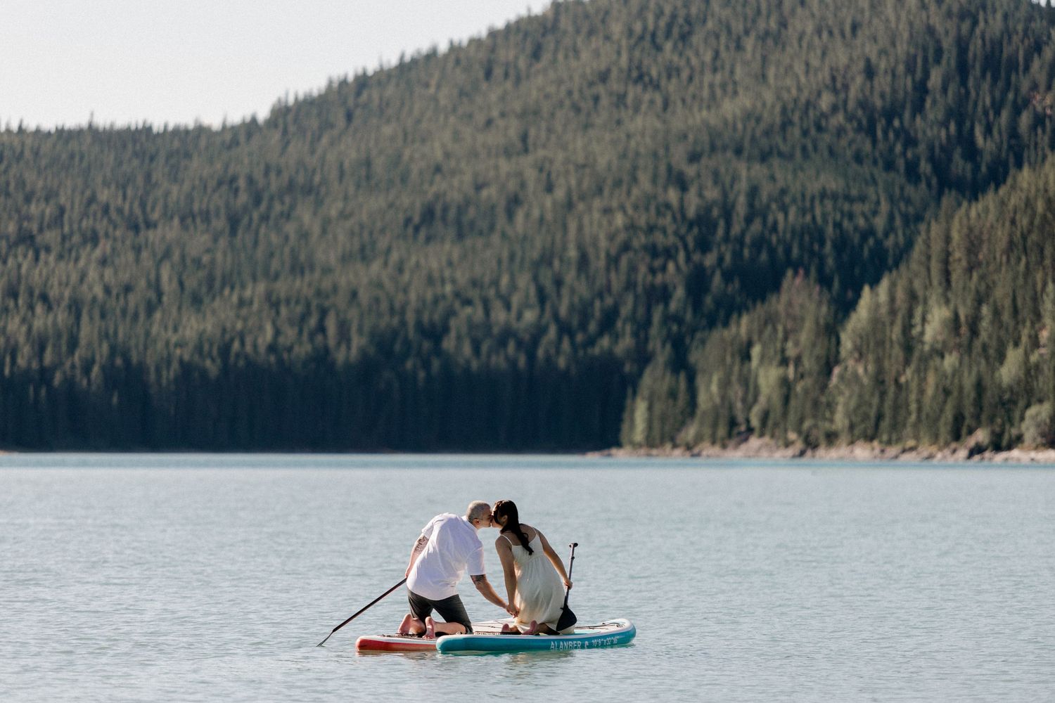 Two people balance on paddleboards on a serene mountain lake surrounded by forest.