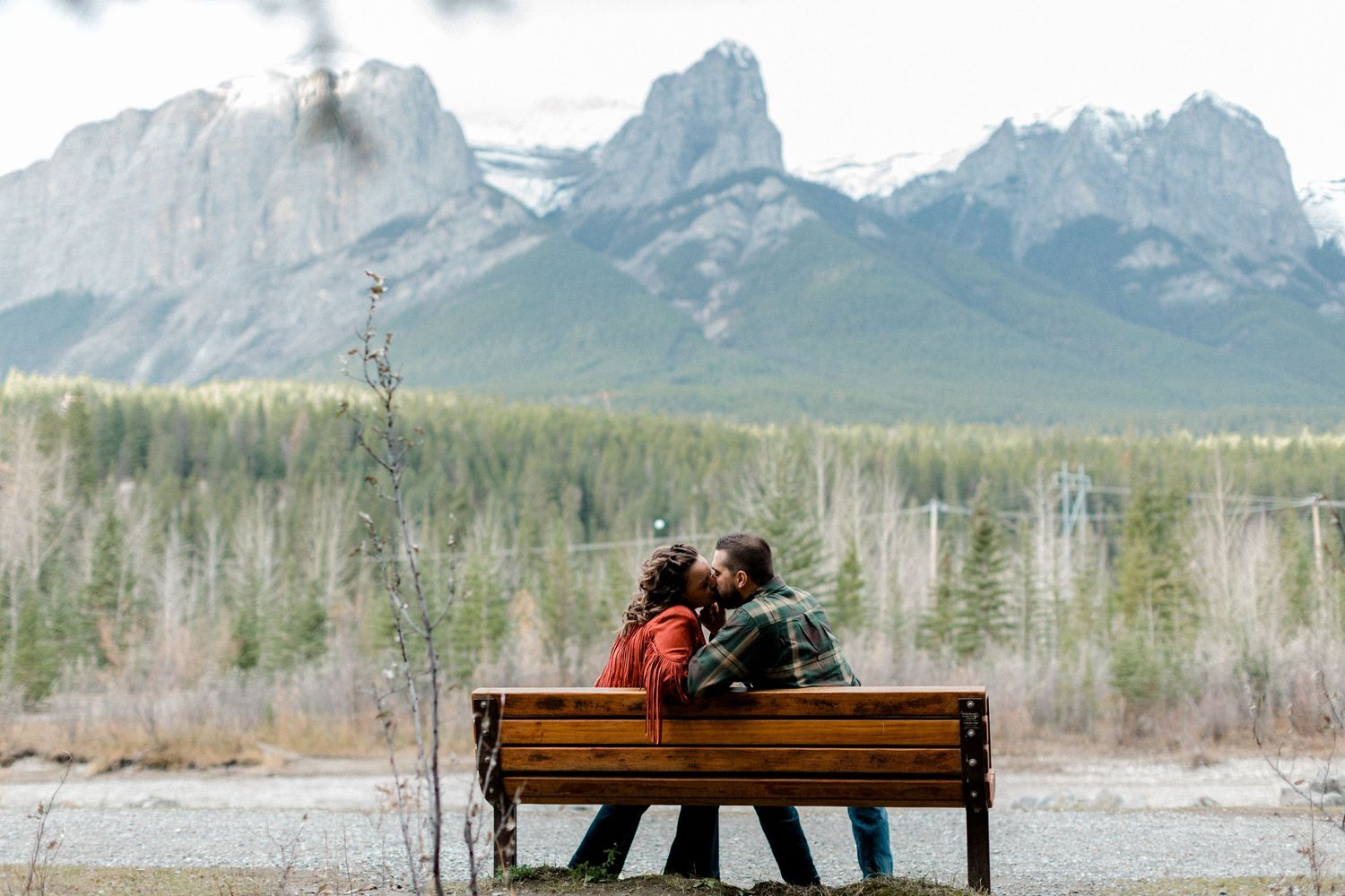 A couple sits on a wooden bench overlooking majestic mountain peaks in the distance.
