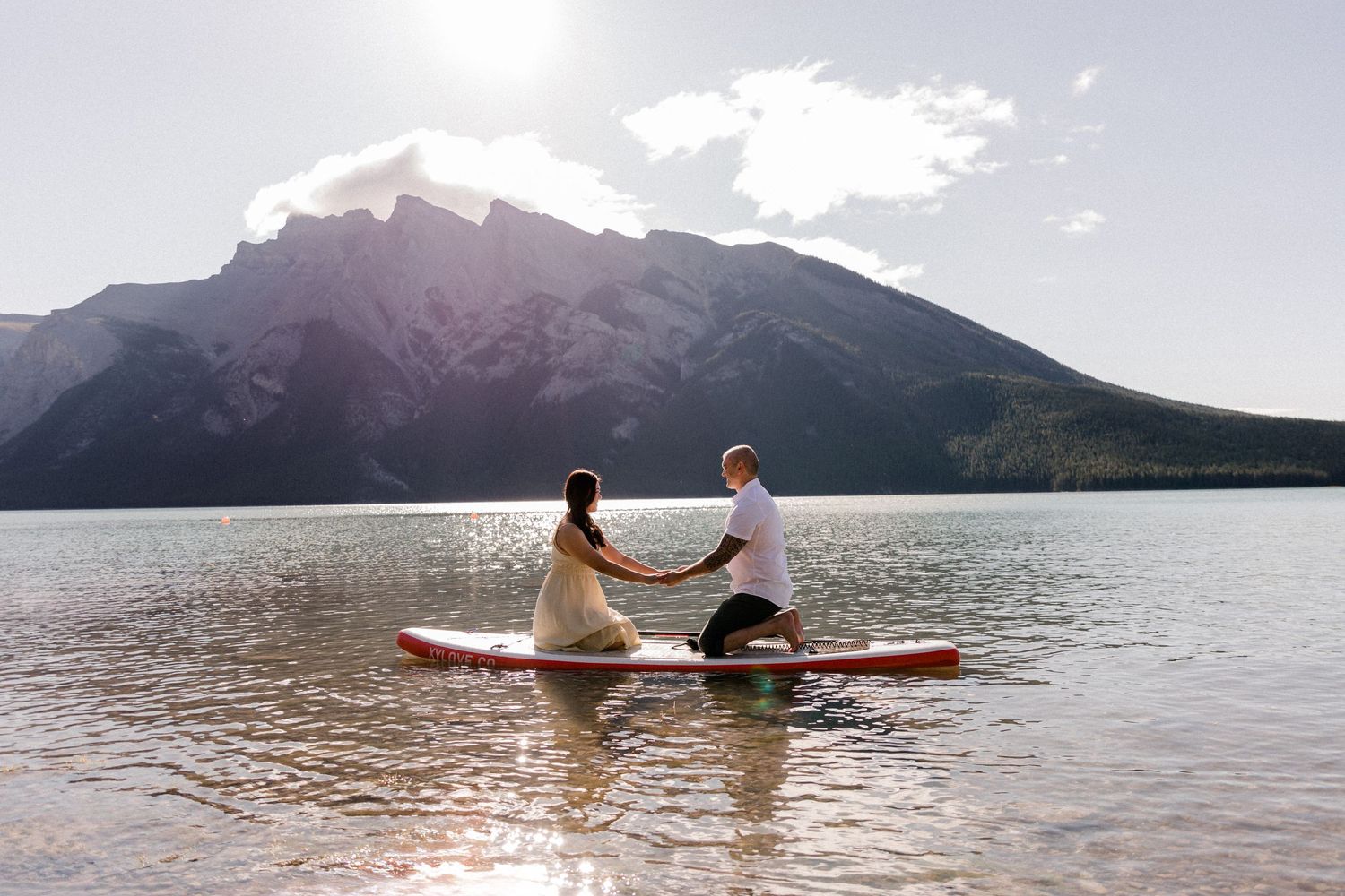 A couple sits together on a paddleboard in a scenic mountain lake on a sunny day.