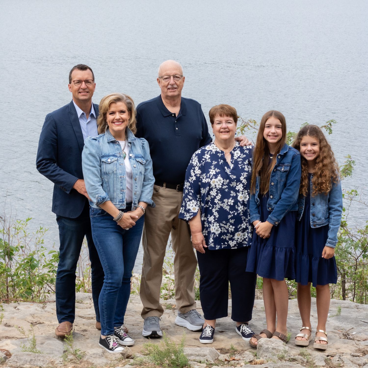 Family with granparents, partents and kids smiling at the camera in front of the bushes and lake.