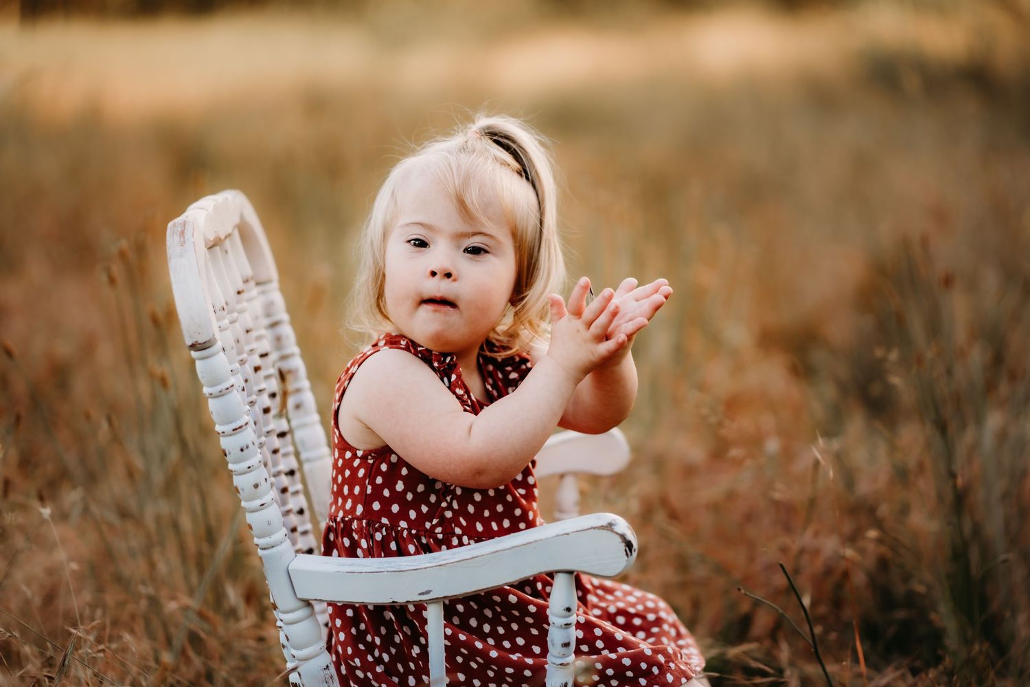 A small child in a red polka dot dress sits in a white vintage chair outdoors during golden hour.
