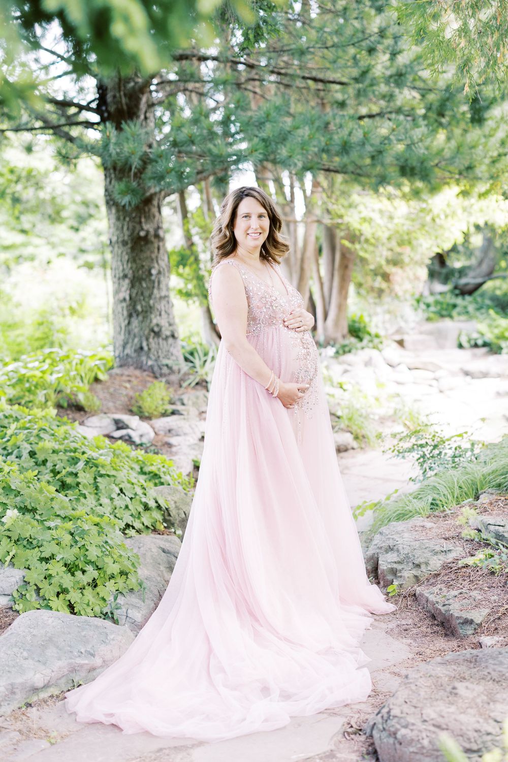 Expectant mother in flowing pink maternity dress holds baby bump in lush garden setting.