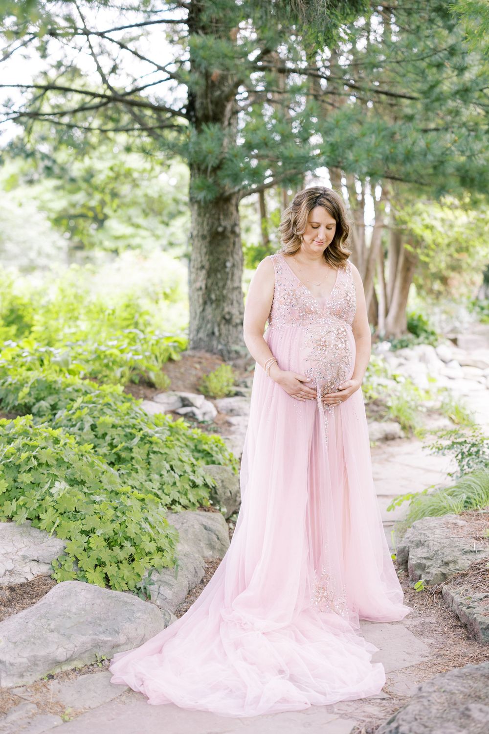 Glowing pregnant woman in pink lace maternity dress looking down at baby bump surrounded by greenery.