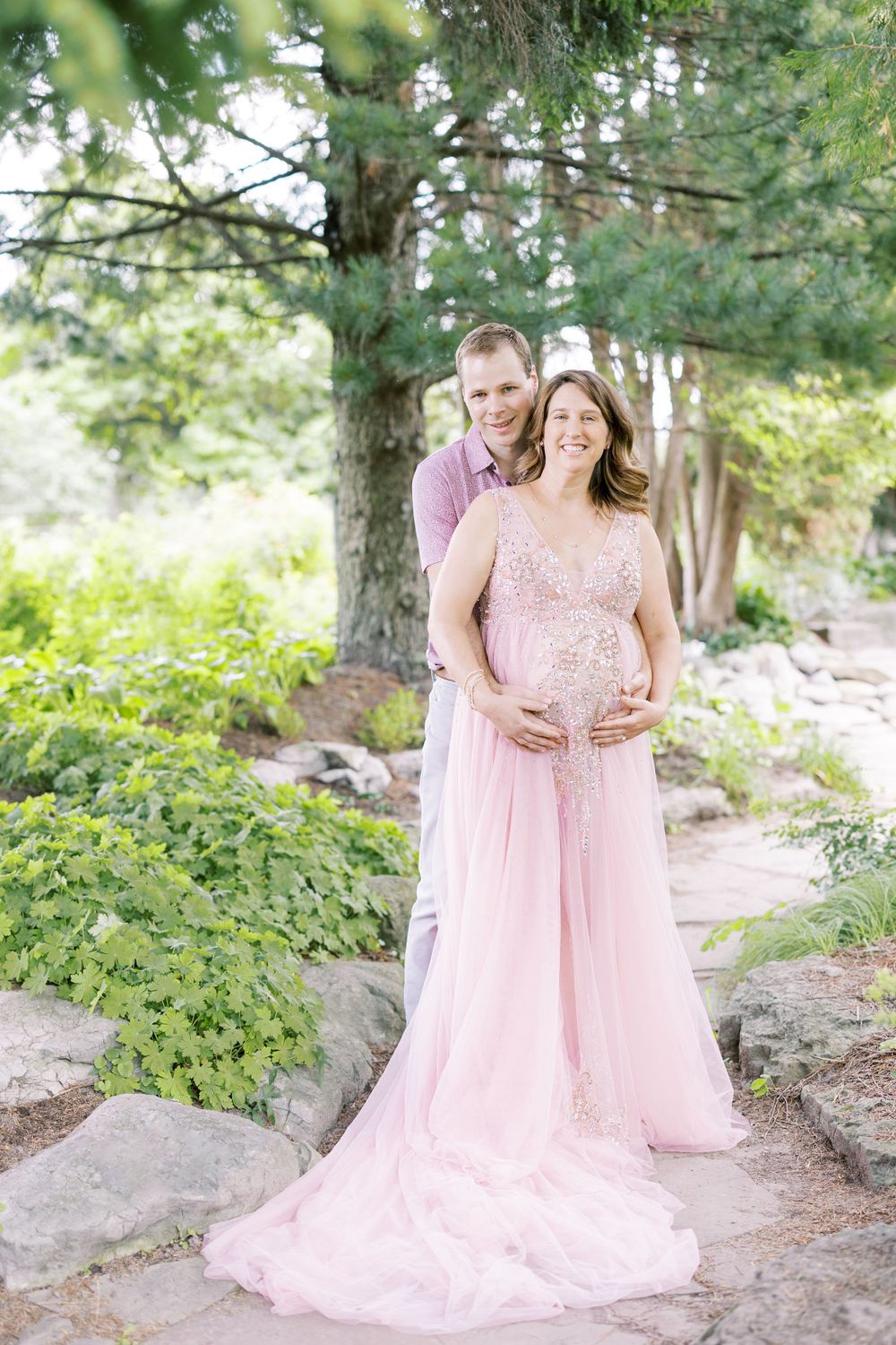 Smiling expectant couple posing together outdoors, man embracing pregnant woman in pink maternity gown.