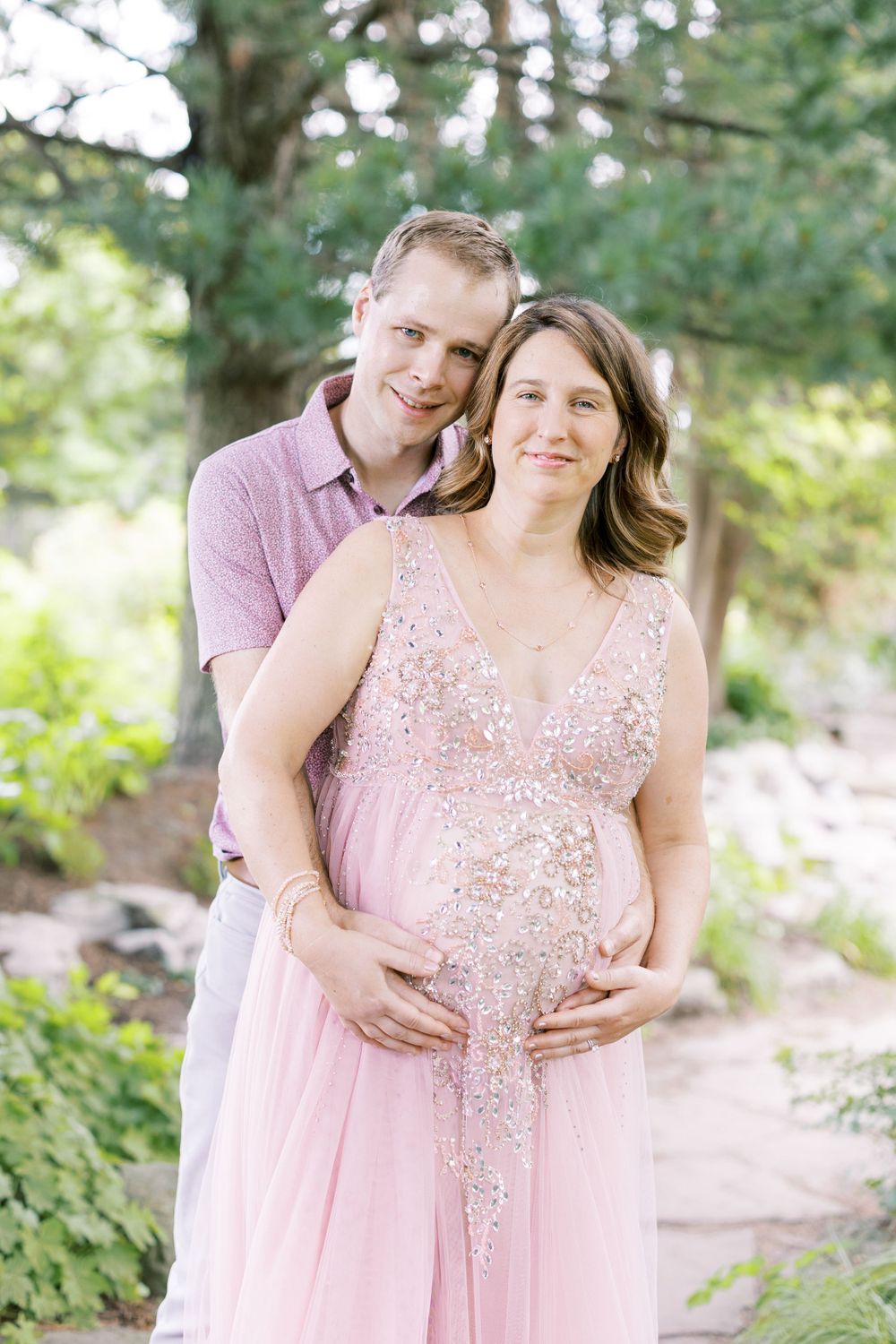 Happy couple posing together, man holding pregnant woman's baby bump, both smiling in garden setting.