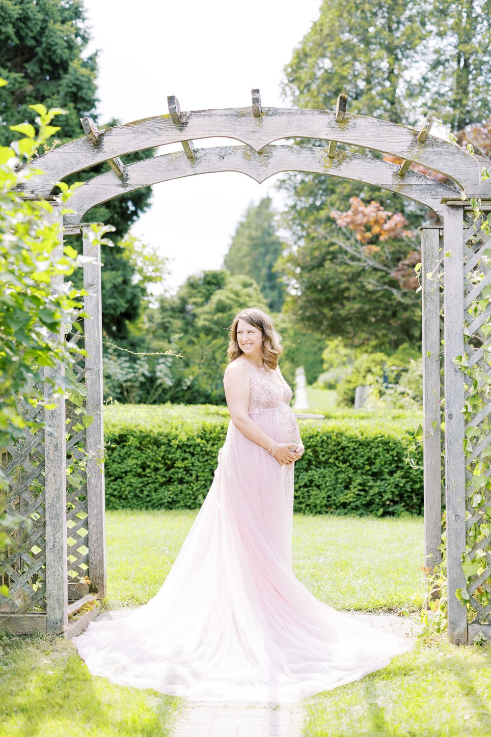 Pregnant woman in flowing pink maternity gown standing gracefully under a rustic wooden garden arbor.