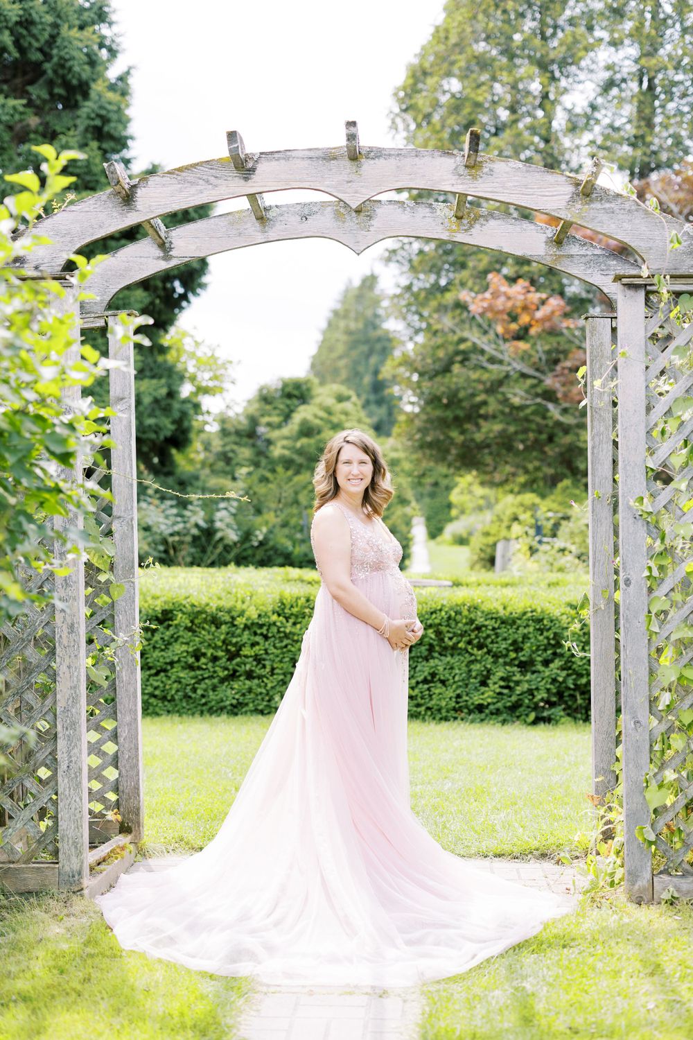 Expectant mother in elegant pink tulle gown posing under weathered wooden arbor in manicured garden.