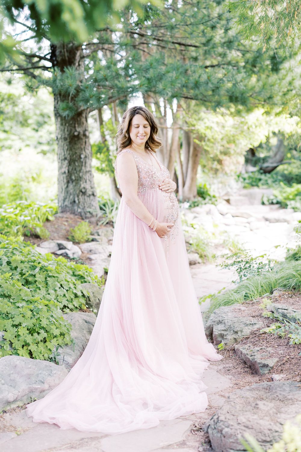 Smiling pregnant woman in blush pink chiffon gown stands among rocks and greenery.