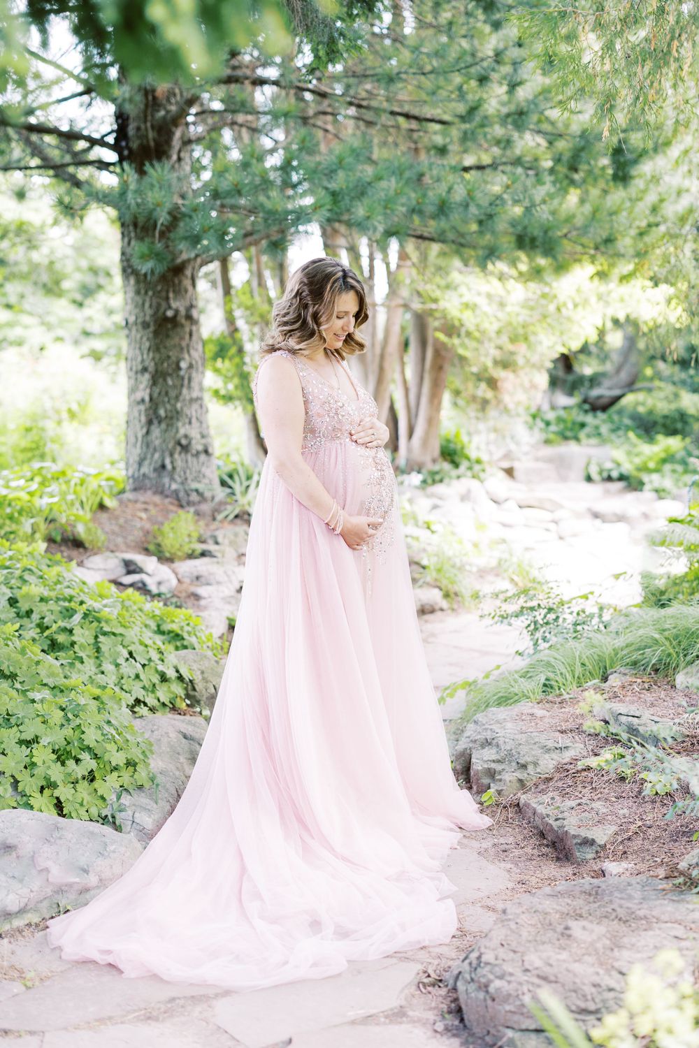 Pregnant woman in elegant pink chiffon gown looks down tenderly at her baby bump.