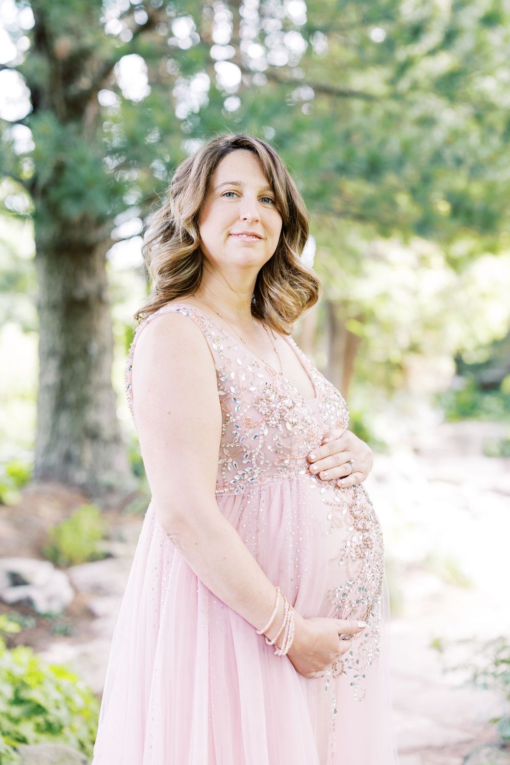 Close-up of pregnant woman in beaded blush pink gown cradling her baby bump outdoors.