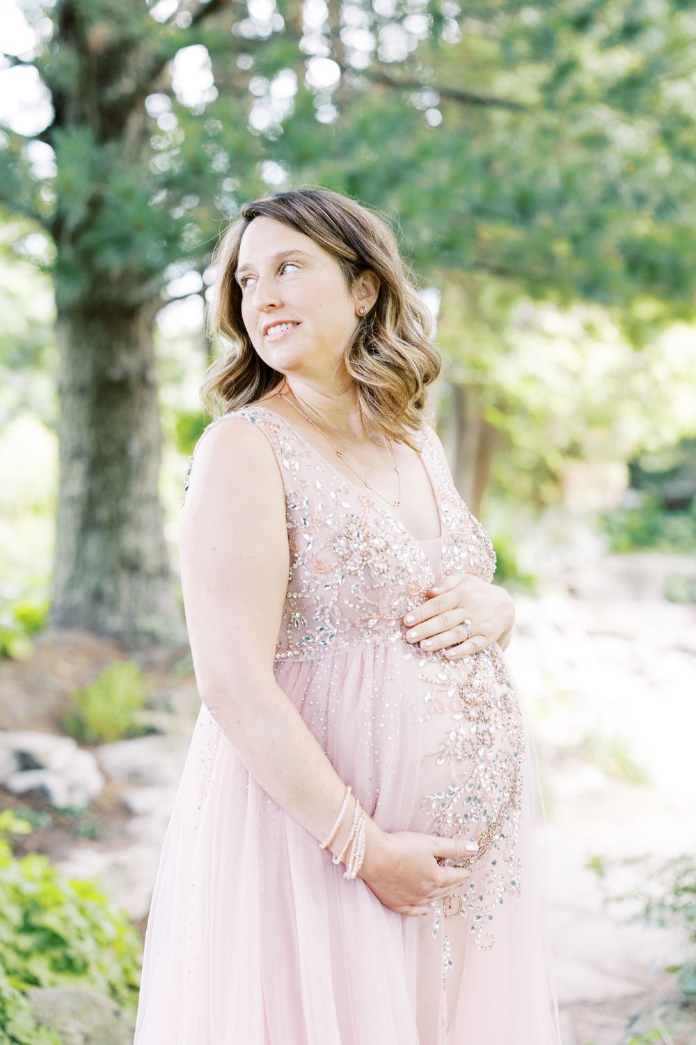 Expectant mother in embellished pink maternity dress gazes softly in sunlit garden setting.