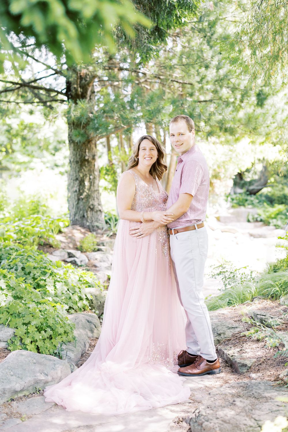 Smiling couple poses together in garden, woman wearing flowing pink beaded maternity gown.