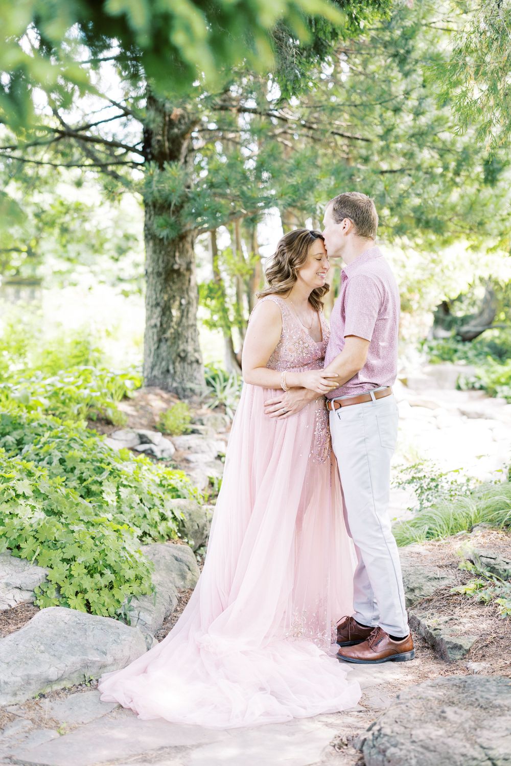 Couple sharing a tender kiss in a lush garden, woman in flowing pink maternity gown, man in pink shirt.