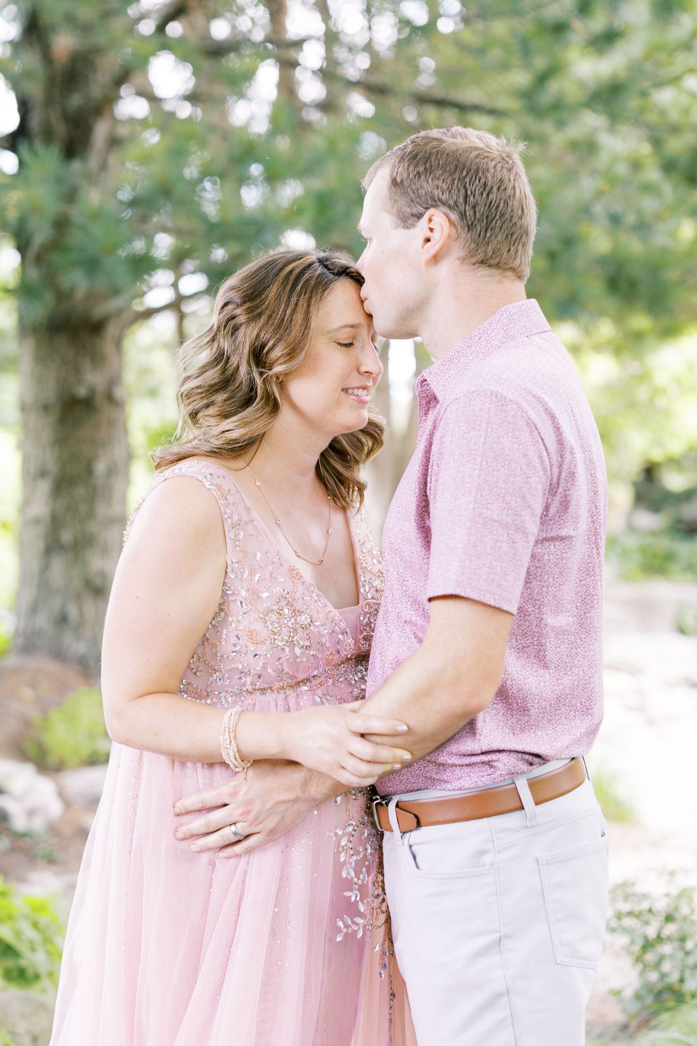 Expectant couple embracing outdoors, man kissing woman's forehead, both in pink tones amid green trees.