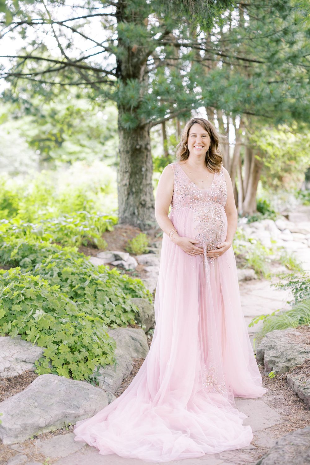 Pregnant woman in elegant pink tulle maternity gown smiling while cradling baby bump in garden setting.