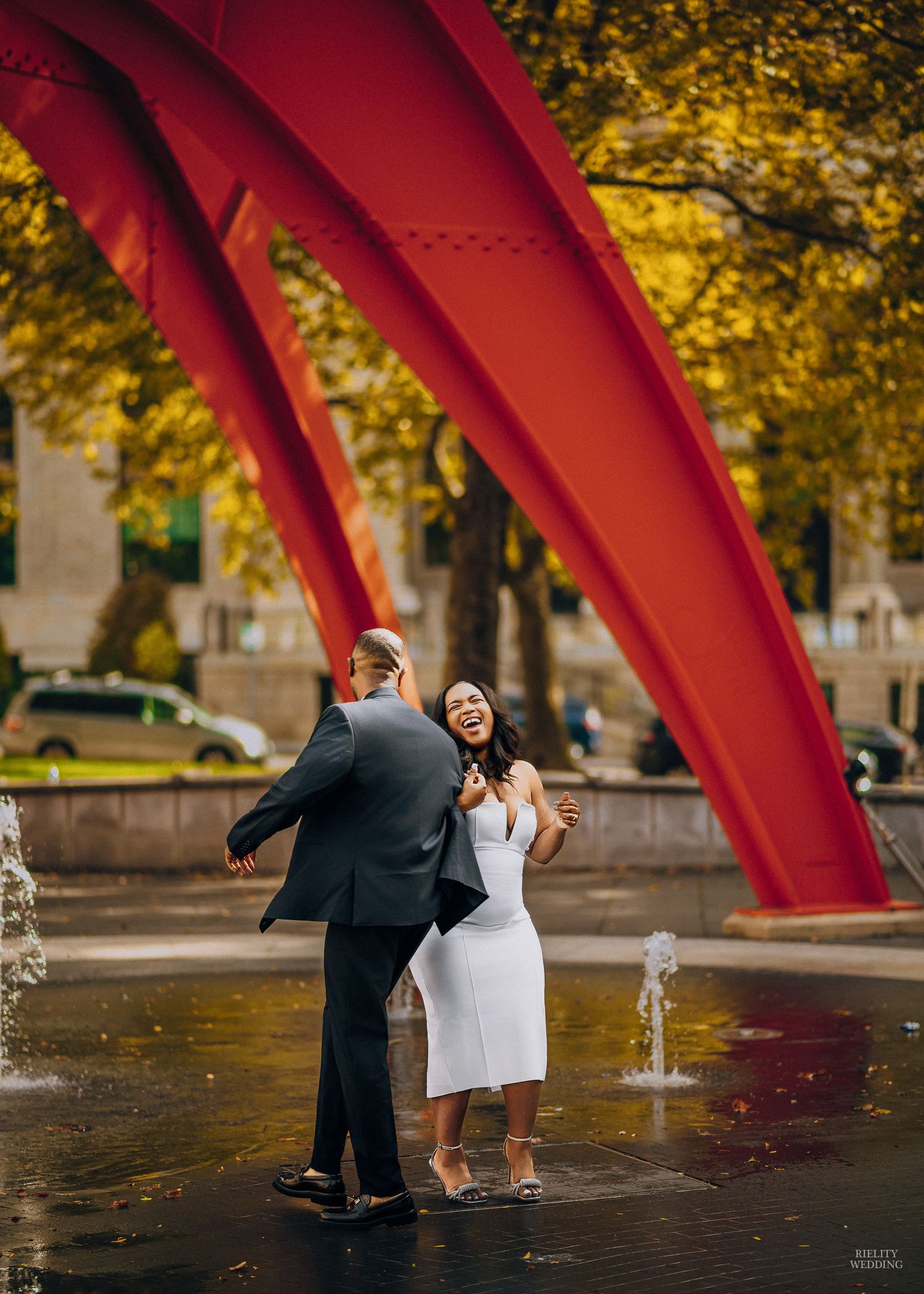 A couple poses playfully near a large red sculptural arch in an urban park setting during autumn.