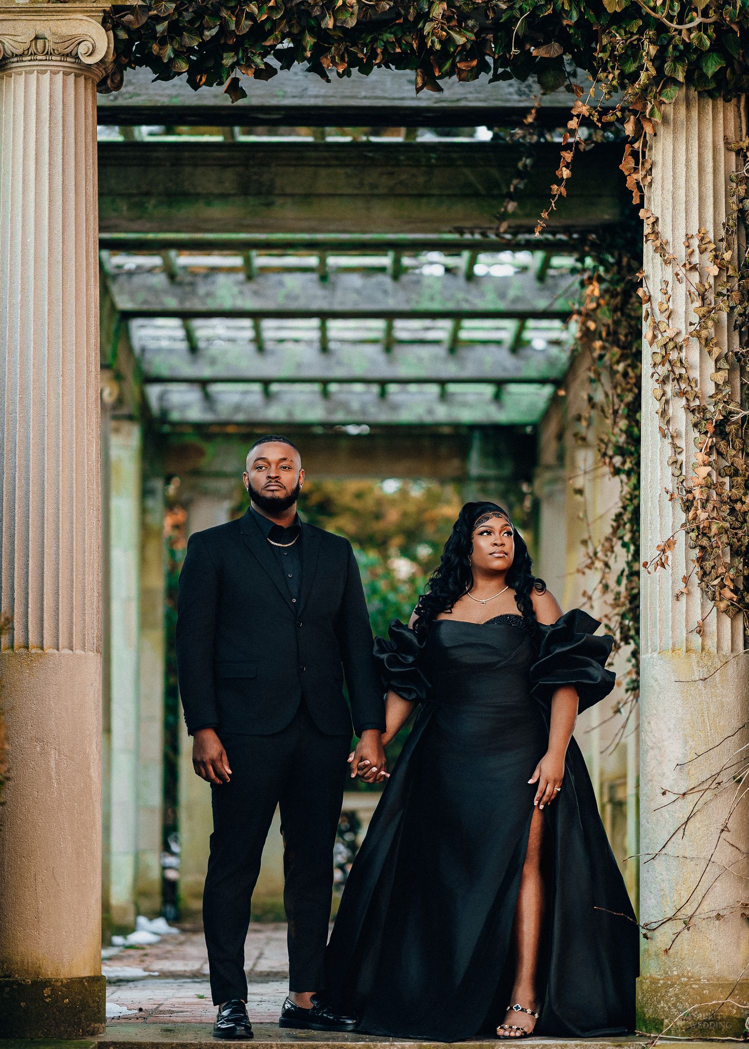 Elegant couple in all black attire pose dramatically between classic stone columns in an outdoor setting.
