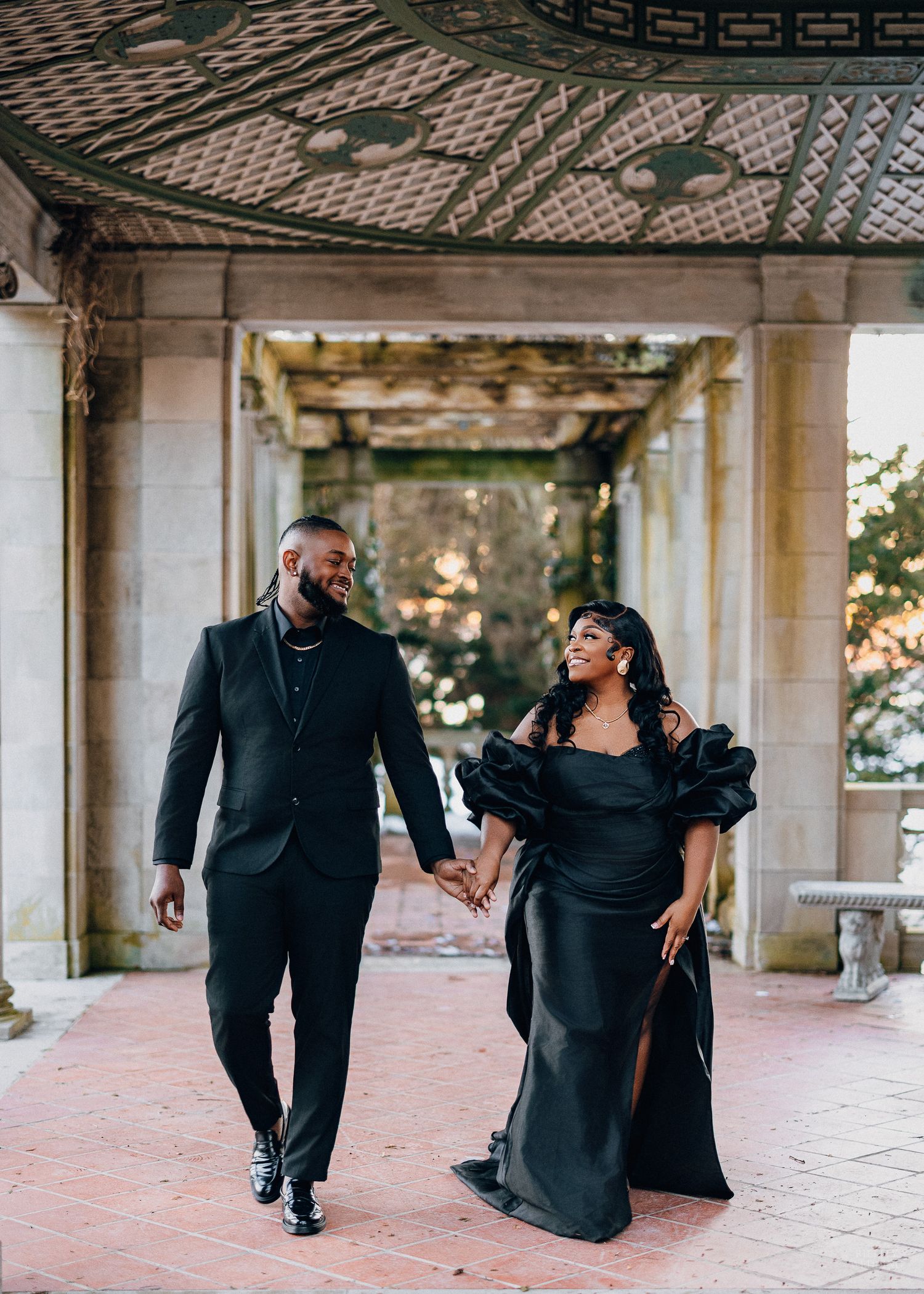 Couple in black formal attire share playful moments while walking through a columned outdoor gallery at sunset.