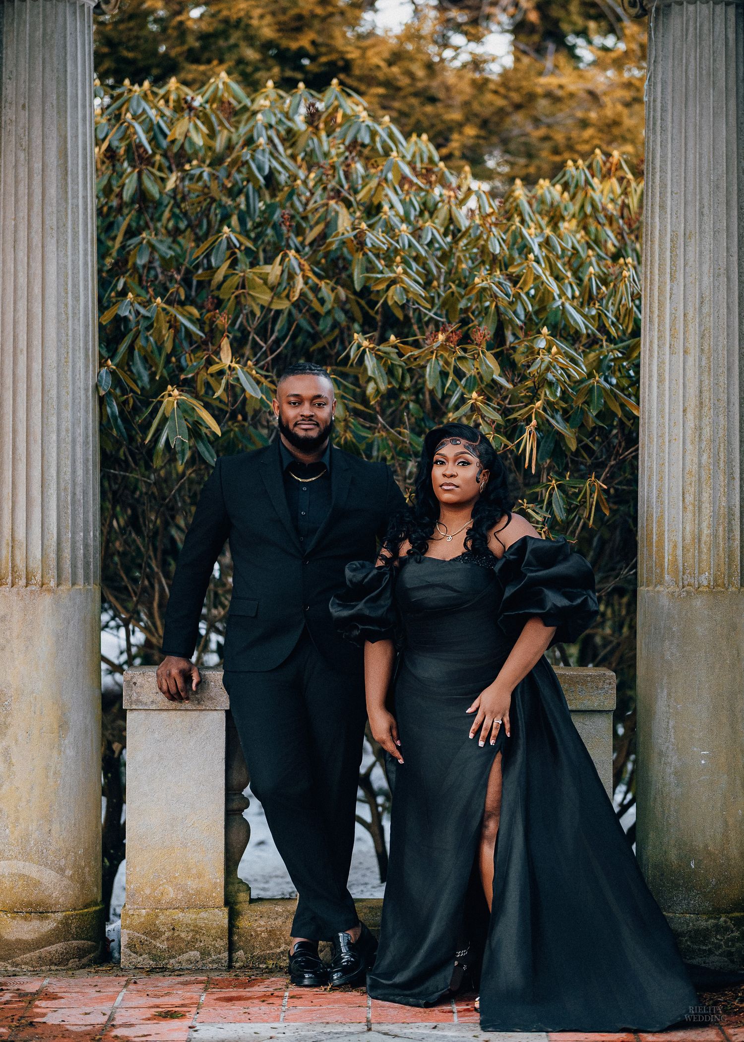 Stylish couple in matching black outfits pose together among classical stone columns surrounded by autumn foliage.