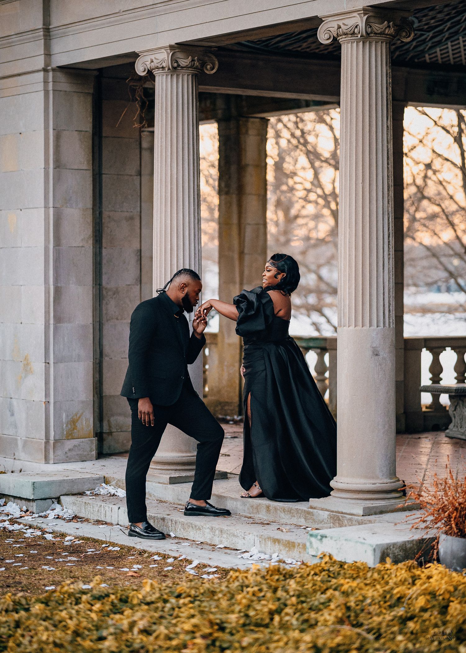 Romantic couple in black outfits pose playfully among classical stone columns with autumn leaves on the ground.
