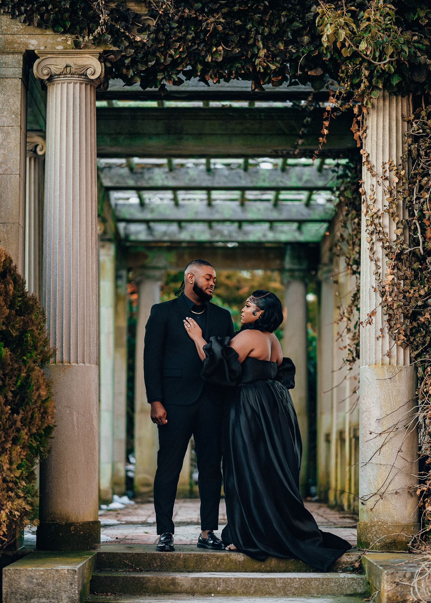 Romantic couple in black formal wear share intimate moments between ornate stone pillars at dusk.