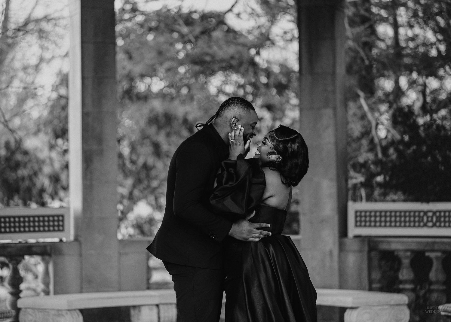 Two figures in dark clothing embrace and dance together on a porch in a black and white photograph.