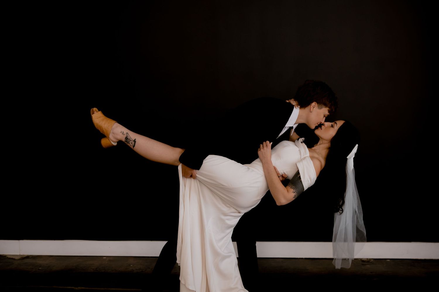 Romantic dance dip pose shows flowing white wedding dress against dark background.