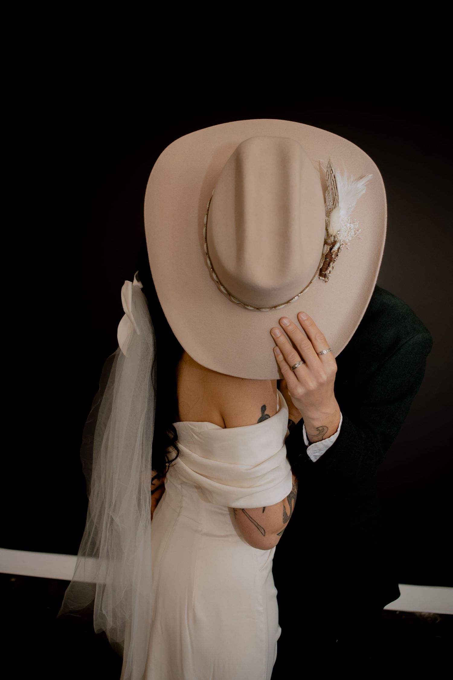Artistic wedding photo with cream cowboy hat and flowing white veil against dark background.