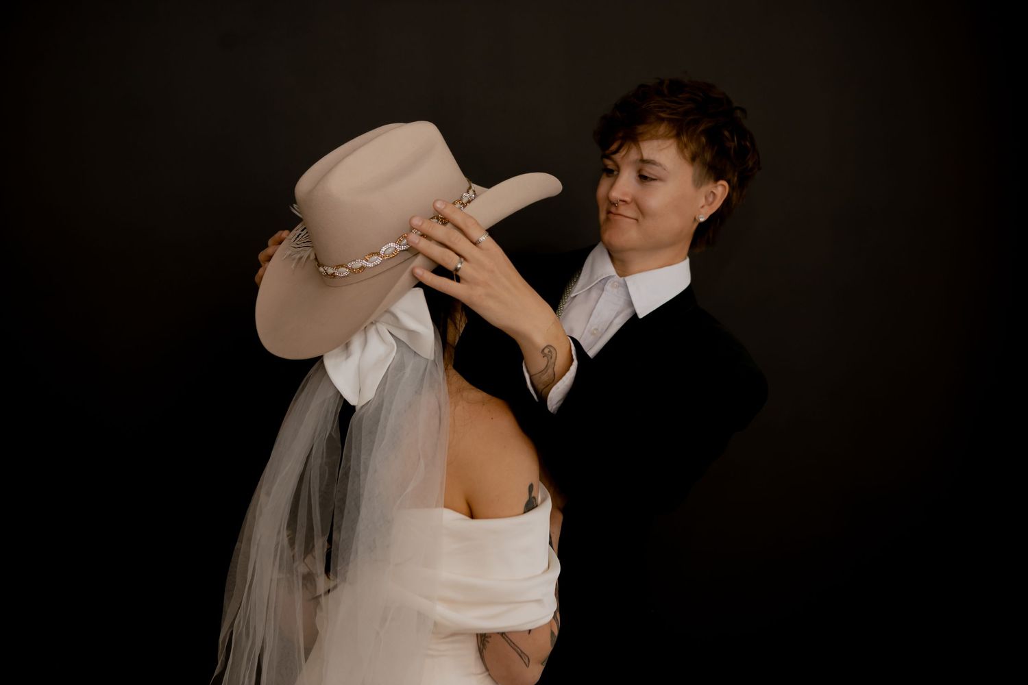 Playful moment with cream cowboy hat and wedding attire against dark backdrop.