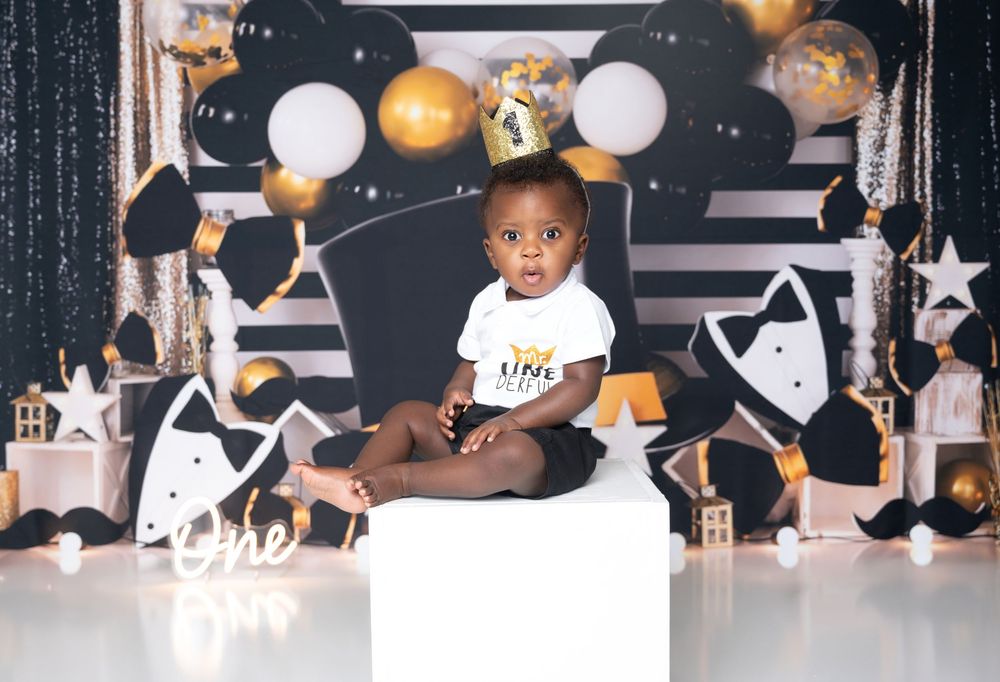 Child sits on white block against backdrop decorated with black and gold balloons, stars and geometric patterns.