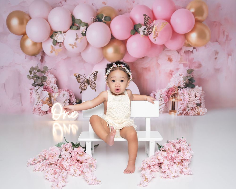 Festive pink and gold balloon arch decorates a studio setup with white bench and floral arrangements.