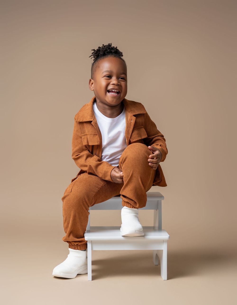Smiling young Black boy in brown corduroy outfit and white boots sitting on a white step stool against beige background.